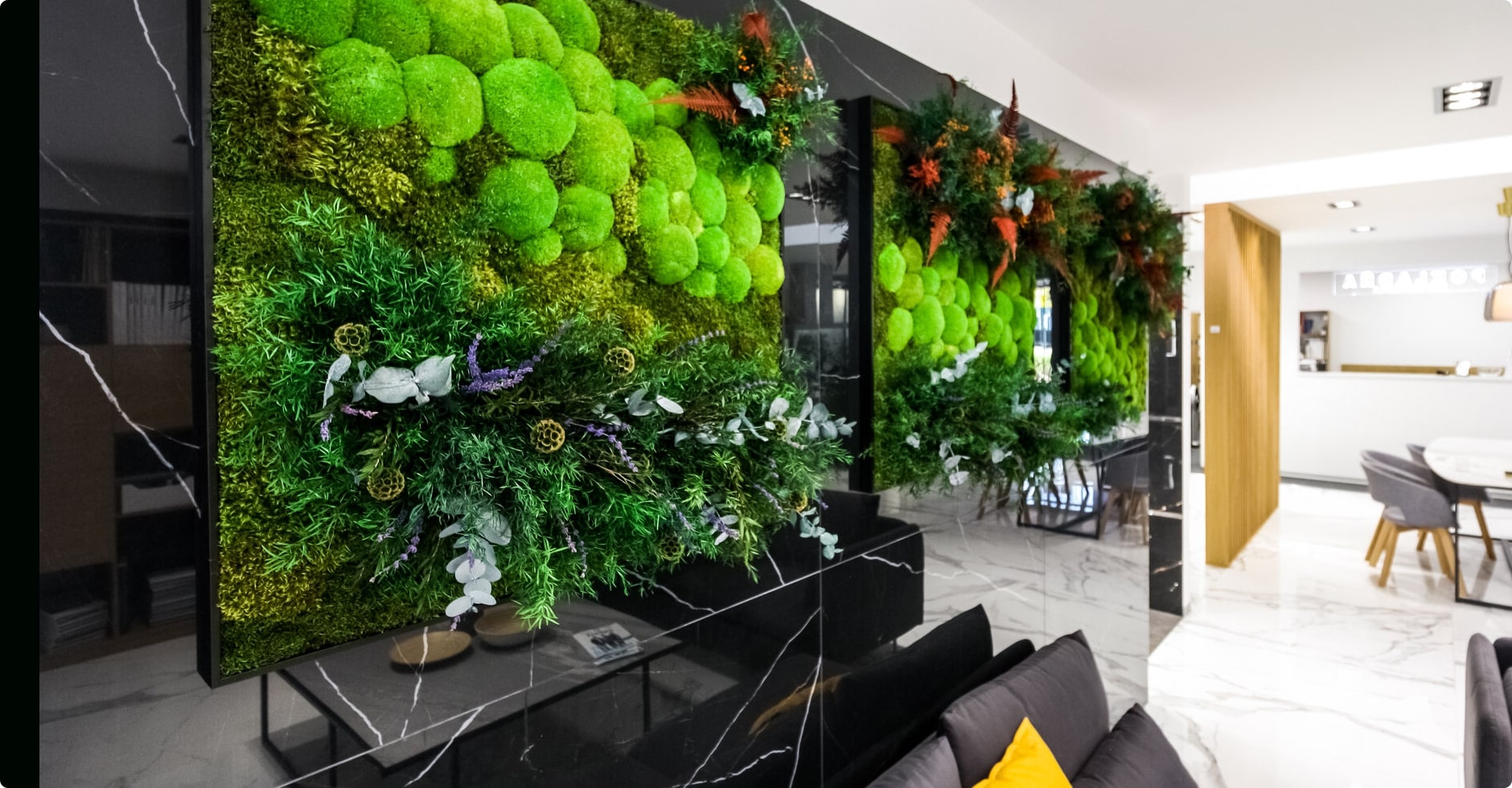 Un intérieur moderne avec des murs ornés de Murs végétaux verts luxuriants avec de la mousse, des plantes et des fleurs. La pièce est décorée de surfaces en marbre noir, d'un canapé gris avec des coussins jaunes et d'un coin repas en arrière-plan avec des chaises blanches et une table en bois.