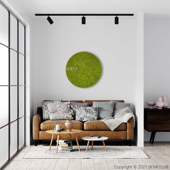 Un salon moderne dispose d'un canapé en cuir marron orné de divers oreillers gris et beiges. Au-dessus du canapé se trouve une décoration murale circulaire en mousse verte réalisée par Tableau de mousse rond en mousse de RENNE mono. Une table basse en bois décorée se trouve devant le canapé et une grande fenêtre permet à la lumière naturelle d'inonder l'espace.