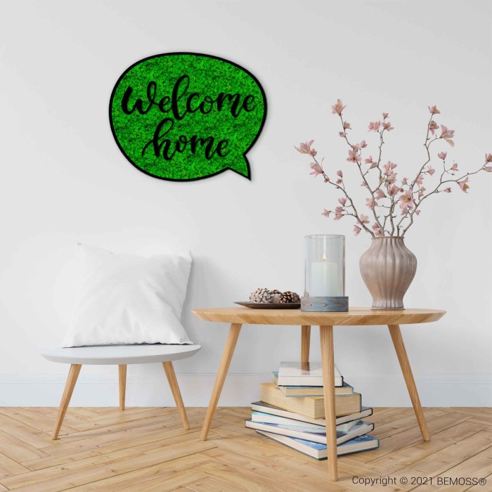 Un coin confortable d'une pièce avec une table d'appoint en bois ornée d'un bonsaï en pot en fleurs, d'une bougie et de pommes de pin décoratives. Une chaise blanche rembourrée se trouve en face et un panneau vert sur le mur blanc indique « Bienvenue à la maison ». L'espace est accentué par le Tableau de mousse Acacia (114x188cm), renforçant la tranquillité. Le sol est en bois clair.