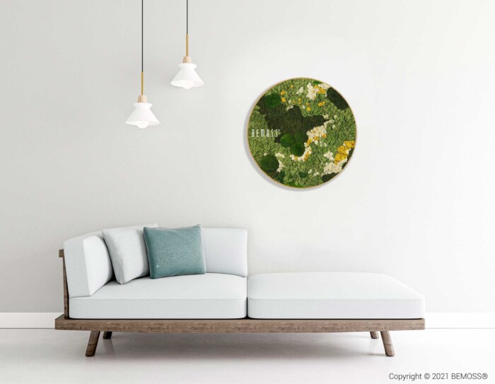 Un espace de vie minimaliste comprend un canapé moderne gris clair avec une structure en bois, orné de coussins verts et gris. Au-dessus, deux suspensions sont suspendues au plafond. Un Tableau de mousse rond BEMOSS® ORTHO ICE rond, vert et jaune, décore le mur blanc derrière le canapé.
