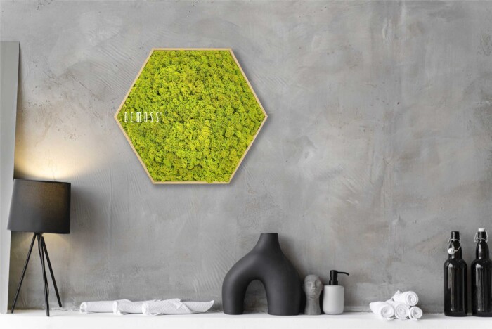 Une salle de bains moderne et minimaliste présente une œuvre d'art hexagonale en mousse verte BEMOSS sur un mur de béton gris. Ci-dessous, il y a une lampe de table noire, un vase sculptural noir, un distributeur de savon, des serviettes roulées blanches et deux bouteilles noires, le tout posé sur une étagère blanche.