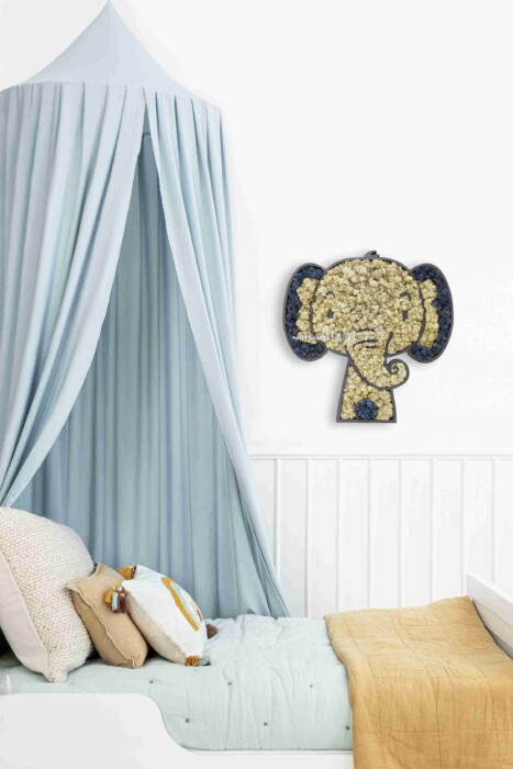 Un lit d'enfant douillet, avec une couverture jaune, des oreillers et un coussin animalier, est installé sous un ciel de lit bleu clair. La tenture murale décorative ÉLÉPHANT est exposée sur le mur blanc au-dessus du lit.
