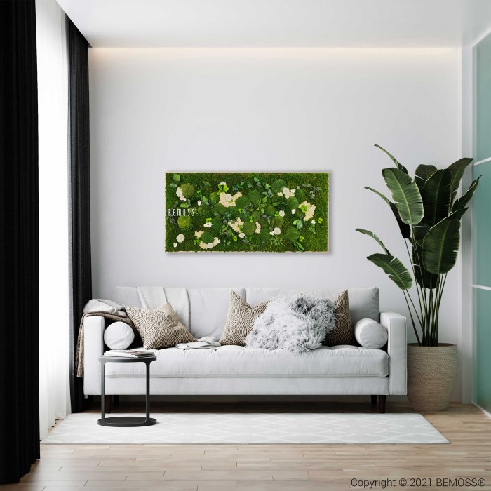 Salon moderne avec un canapé gris clair orné de coussins douillets et d'un jeté moelleux. Une petite table d'appoint noire à côté du canapé contient une tasse. Le mur présente une œuvre d’art rectangulaire Tableau de mousse PLANTES Mesia Natural. Une grande plante en pot se dresse dans le coin, ajoutant une touche naturelle.