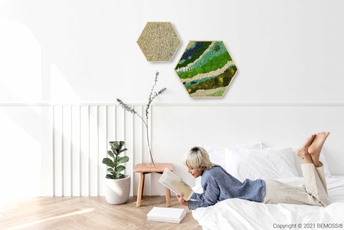 Un enfant aux cheveux blonds courts est allongé sur le ventre sur un lit blanc, lisant un livre. Deux œuvres d'art murales hexagonales sont suspendues au-dessus d'elles : l'une avec un motif beige texturé et l'autre présentant un motif coloré vert et bleu. Une plante en pot et une petite table d'appoint fleurie complètent ce serein Hexagone de mousse BEMOSS® ORTHO BORJA.