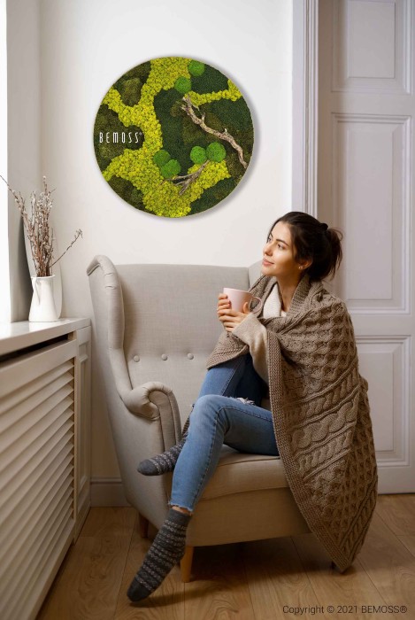 Une femme est assise dans un fauteuil enveloppée dans une couverture tricotée, tenant une tasse. Elle regarde une décoration murale verte vibrante comportant des motifs de mousse et d'arbres avec le texte « BEMOSS » dessus. La pièce a des murs blancs et une lumière naturelle entrant à travers une fenêtre, soulignant la beauté du Tableau de mousse rond BEMOSS® ORTHO BORJA.