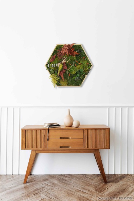 Un intérieur minimaliste comprend un buffet en bois avec des pieds inclinés placés contre un mur blanc avec des détails en panneaux. Au mur, un tableau de mousse avec mousse et feuillage stabilisés est suspendu au-dessus du buffet qui contient deux vases en céramique et un livre fermé.