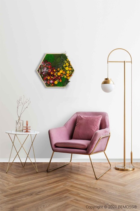 Un intérieur moderne comprend un fauteuil rose moelleux avec des accents dorés, associé à une table d'appoint blanche et dorée contenant une plante et des objets de décoration. Un lampadaire élégant se trouve à proximité. Au-dessus du mur blanc est accroché un tableau de mousse vibrant, ajoutant une touche de nature à l'espace.