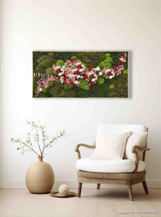 Une scène d'intérieur cosy mettant en scène un fauteuil beige aux coussins blancs placé à côté d'un vase rond en céramique contenant une petite plante aux branches délicates. Au-dessus de la chaise, il y a un Tableau de mousse rectangulaire PLANTES Acebo BLUE avec de la mousse verte et des fleurs roses, rouges et blanches.