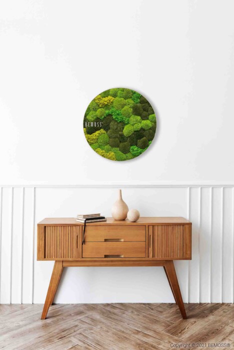 Un buffet en bois avec deux tiroirs et des pieds fuselés se dresse contre un mur blanc. Au-dessus, il y a deux vases beiges et une pile de livres. Au-dessus du buffet, une œuvre d'art murale ronde verte avec différentes nuances de mousse est accrochée au mur lambrissé blanc.