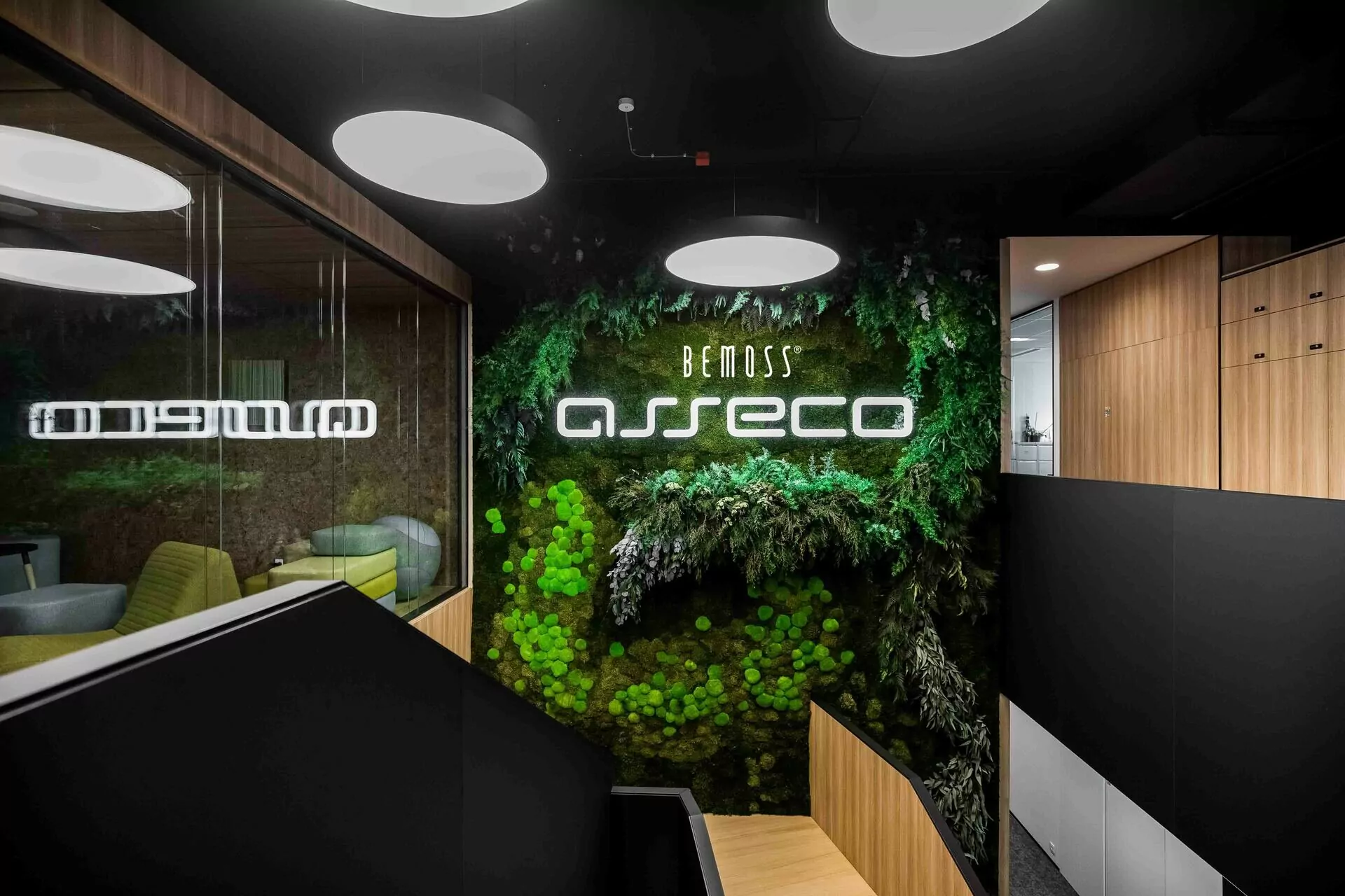 Espace de bureau moderne avec un tableau de mousse vert affichant le texte néon « Bemoss Asseco ». La zone est ornée de plafonniers ronds et de boiseries. Une grande baie vitrée offre une vue sur une pièce meublée avec des chaises de bureau et une table.