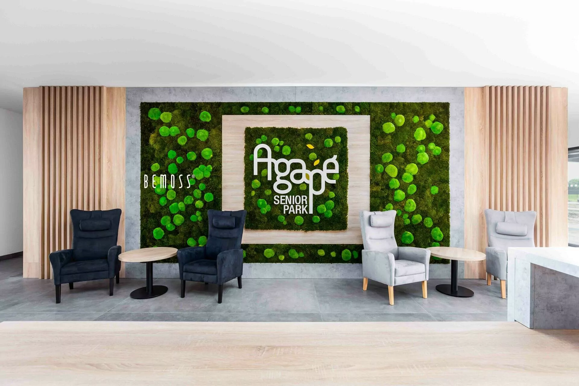Un espace d'attente moderne et élégant comprend des fauteuils confortables et des petites tables rondes. Le mur abrite un jardin vertical vert, connu sous le nom de « Murs végétaux », avec les mots « Agape Senior Park » bien en évidence au centre. Le design est épuré et accueillant.