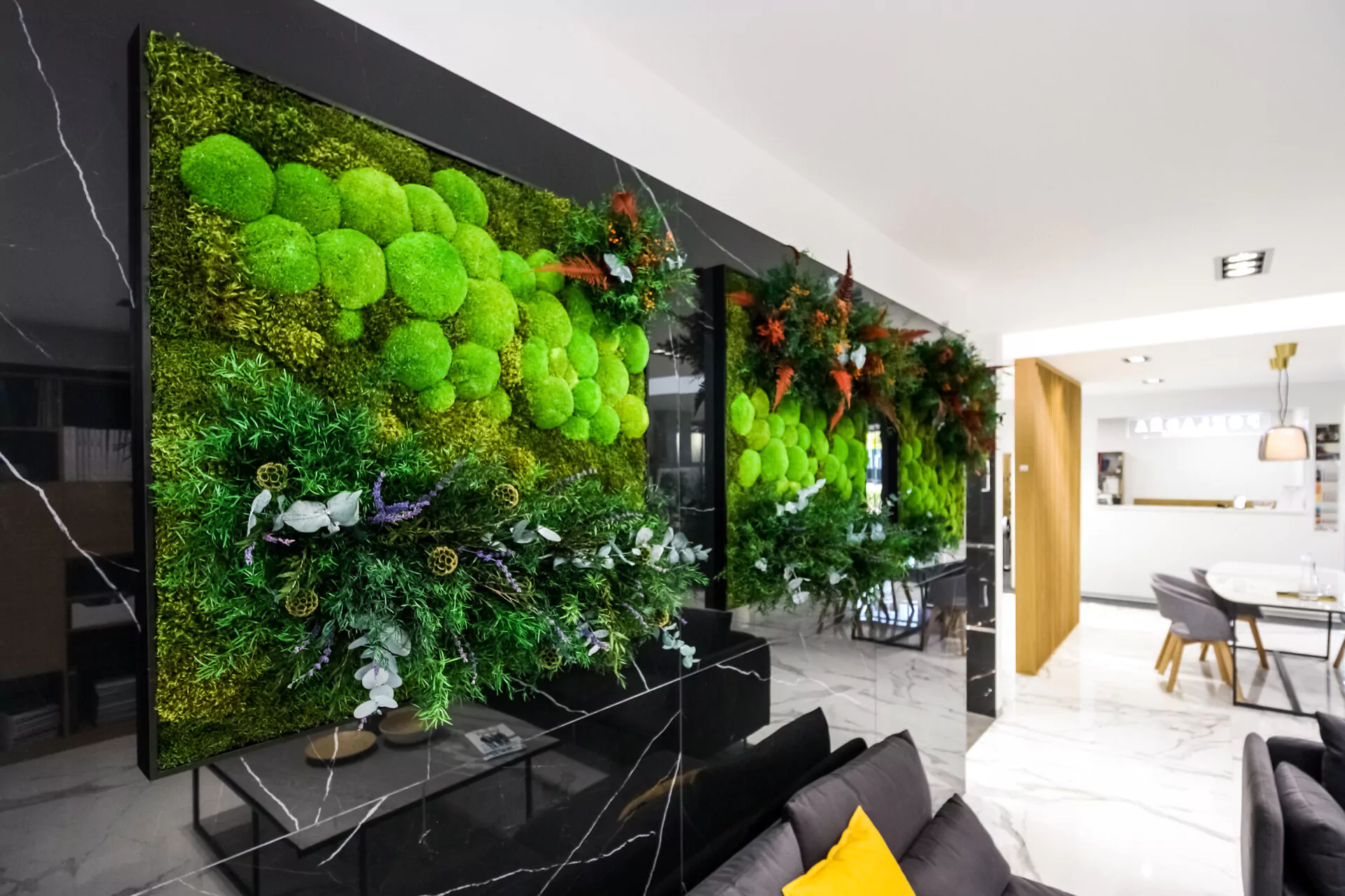 Un salon moderne avec un mur d'accent en marbre noir avec des jardins verticaux luxuriants et un tableau de mousse BEMOSS. La pièce dispose d'un canapé gris avec des oreillers jaunes et gris, du carrelage blanc brillant et une vue dégagée sur la salle à manger au décor minimaliste.