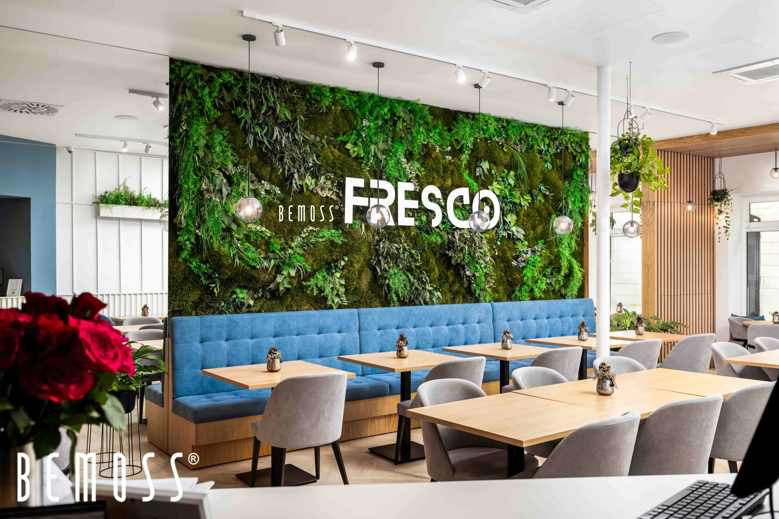 Intérieur de restaurant moderne avec un mur végétal vert portant le mot « FRESCO ». L'espace comprend des banquettes rembourrées bleues, des tables en bois clair et des chaises grises. Les fleurs rouges sont à gauche. La marque « BEMOSS » est visible dans le coin inférieur gauche, complétant le design du tableau de mousse.