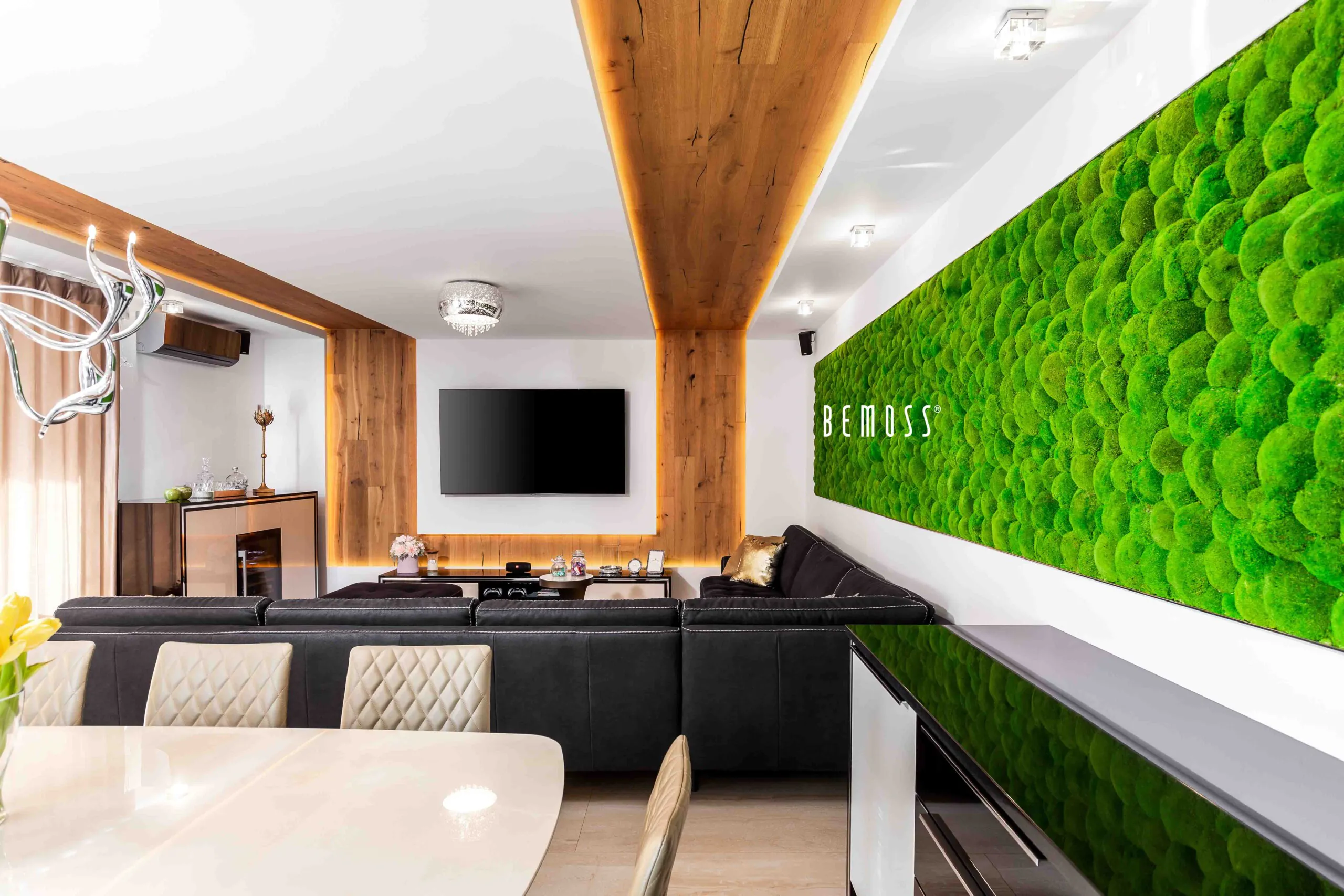 Salon moderne avec poutres apparentes au plafond, grand canapé sectionnel noir et télévision murale à écran plat. Un mur de mousse verte vibrante avec le mot « BEMOSS » est présenté sur la droite. Au premier plan se trouve le coin repas avec des chaises matelassées beiges et une table blanche.