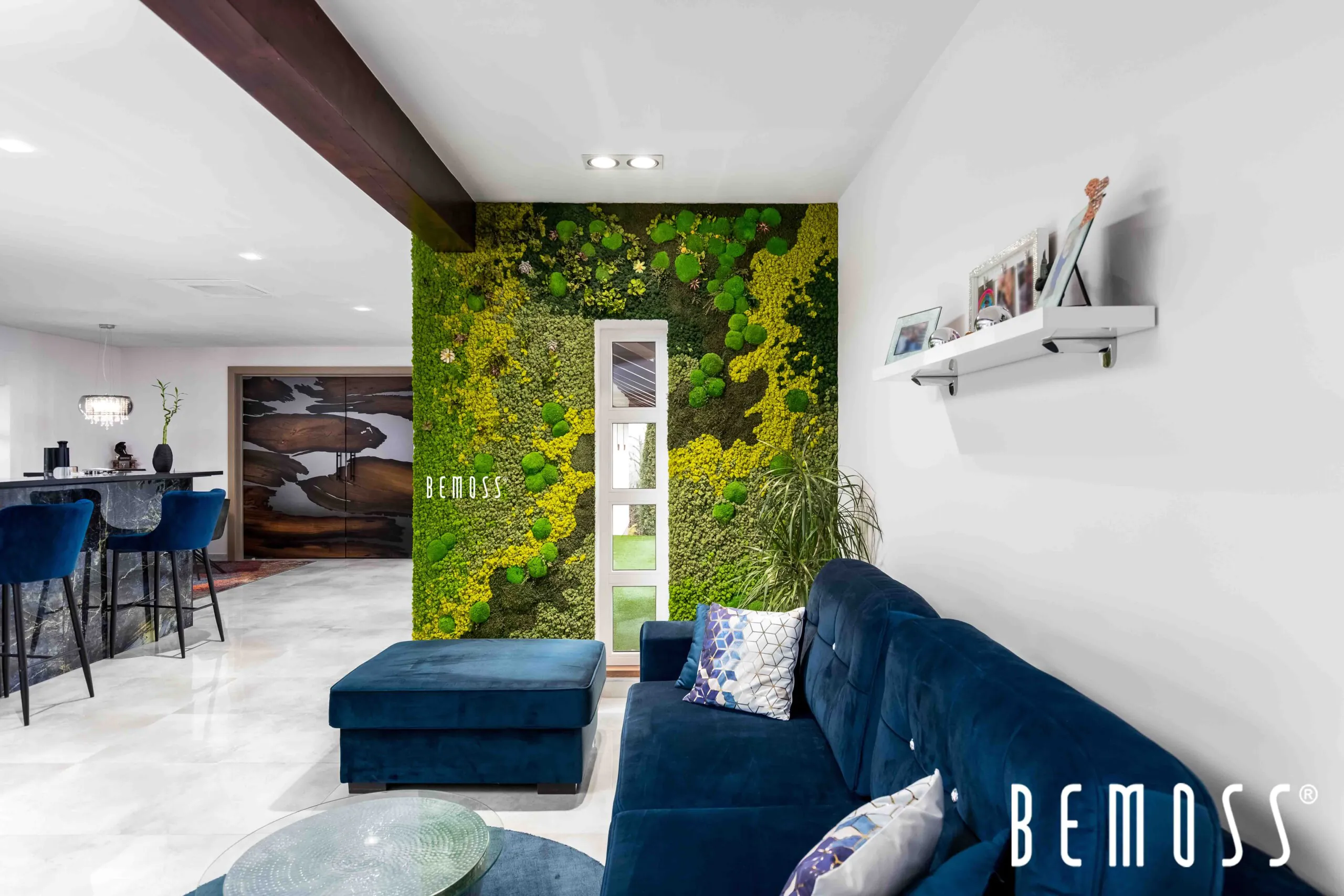 Un salon moderne avec un mur de mousse verte vibrante derrière une porte blanche. La pièce comprend un canapé en velours bleu foncé, un pouf assorti et une table basse en verre. À droite, une étagère blanche contient des objets de décoration. Le logo BEMOSS est visible dans le coin de ce saisissant tableau de mousse.