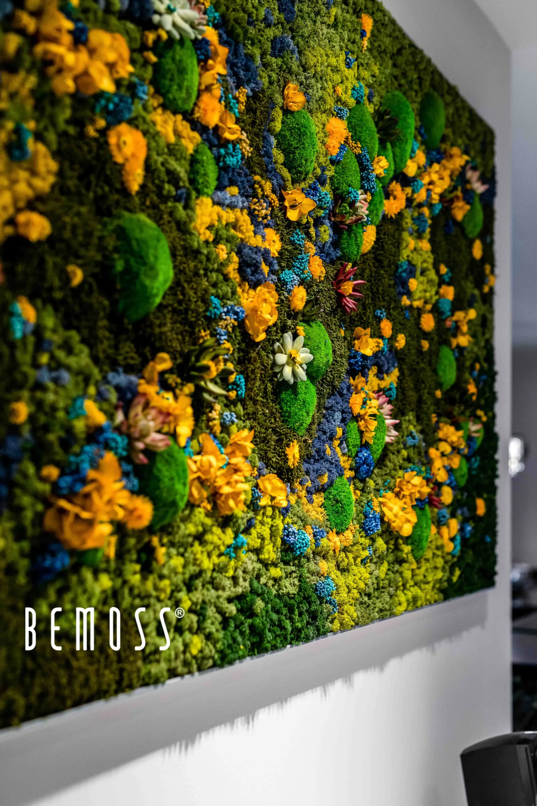 Tenture murale verticale vibrante faite de mousse préservée et de fleurs artificielles, ce tableau de mousse présente un mélange texturé de monticules de mousse verte, de fleurs jaune vif et des notes d'accents bleus et violets, créant un affichage luxuriant et coloré. Le mot « BEMOSS » est visible en bas.