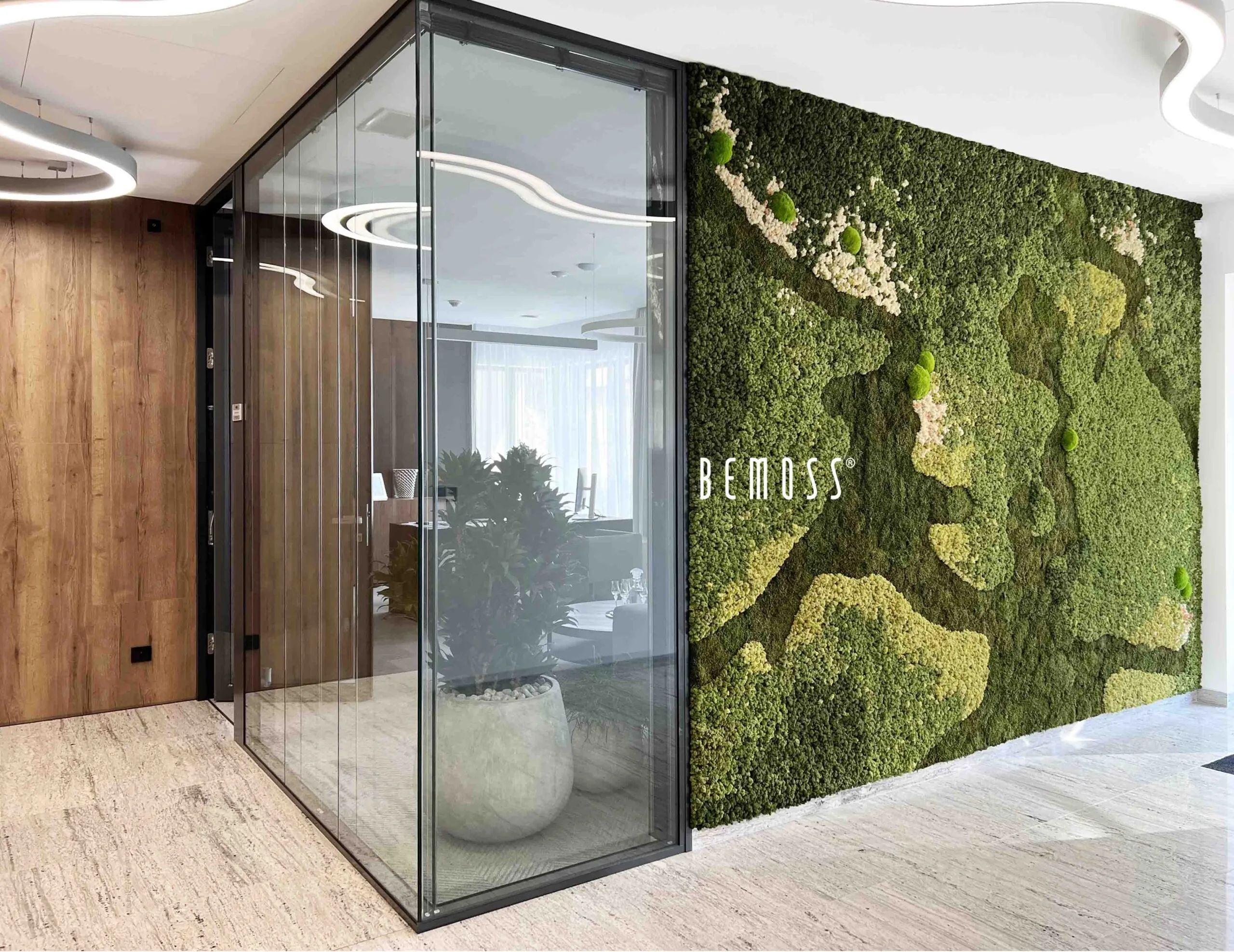 Un espace de bureau moderne comprend une salle de conférence aux parois de verre et un grand mur de mousse verte avec des motifs abstraits sur le côté droit. Le mot « BEMOSS » est affiché sur les murs végétaux. La décoration intérieure comprend des éléments en bois et de couleurs claires.